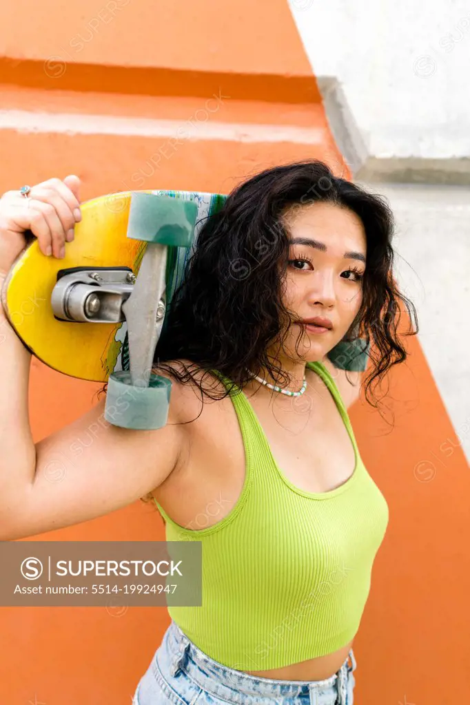 Asian woman standing next to the orange wall holding a skateboar