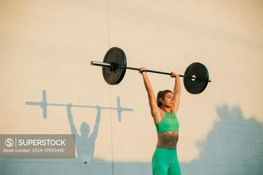 Young active woman doing overhead press with barbell in morning 