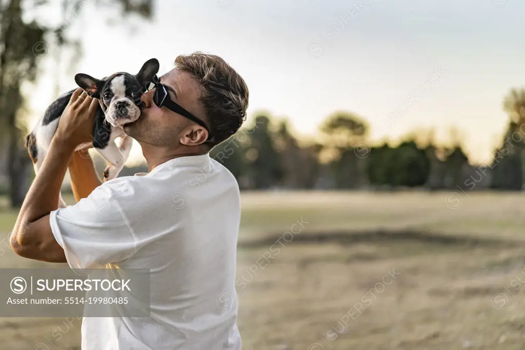 Handsome young man holding up and kissing a french bulldog.