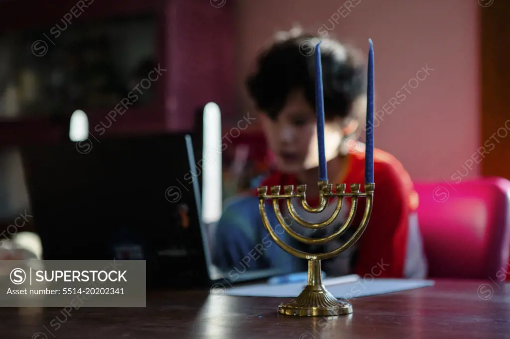 A boy sits at table on a computer, a menorah in foreground
