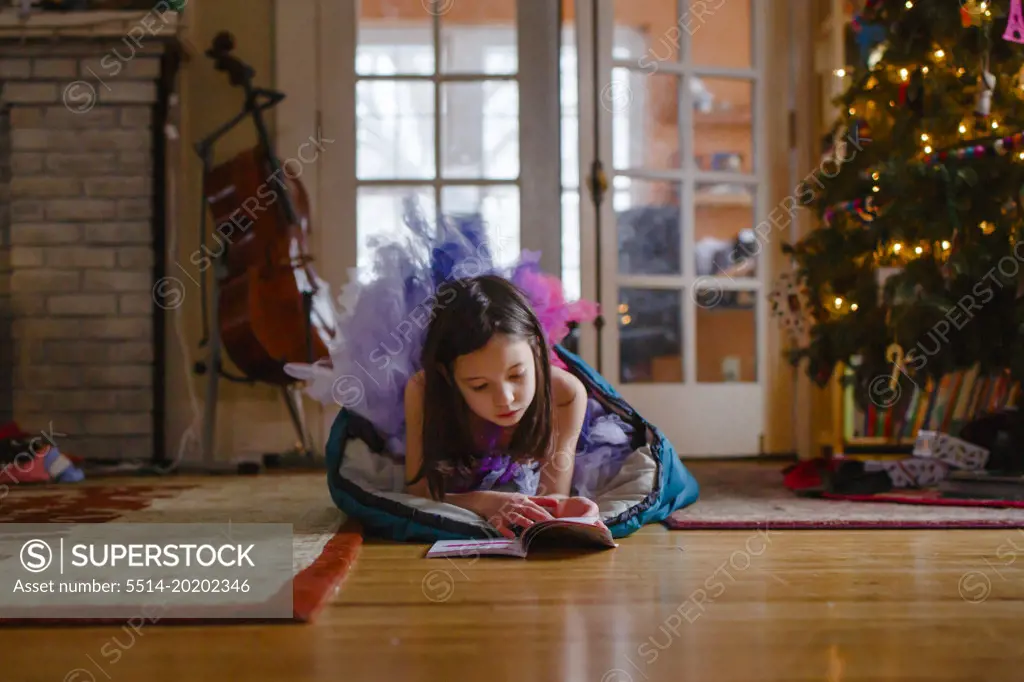 A small girl in tutu reads on floor by Christmas tree