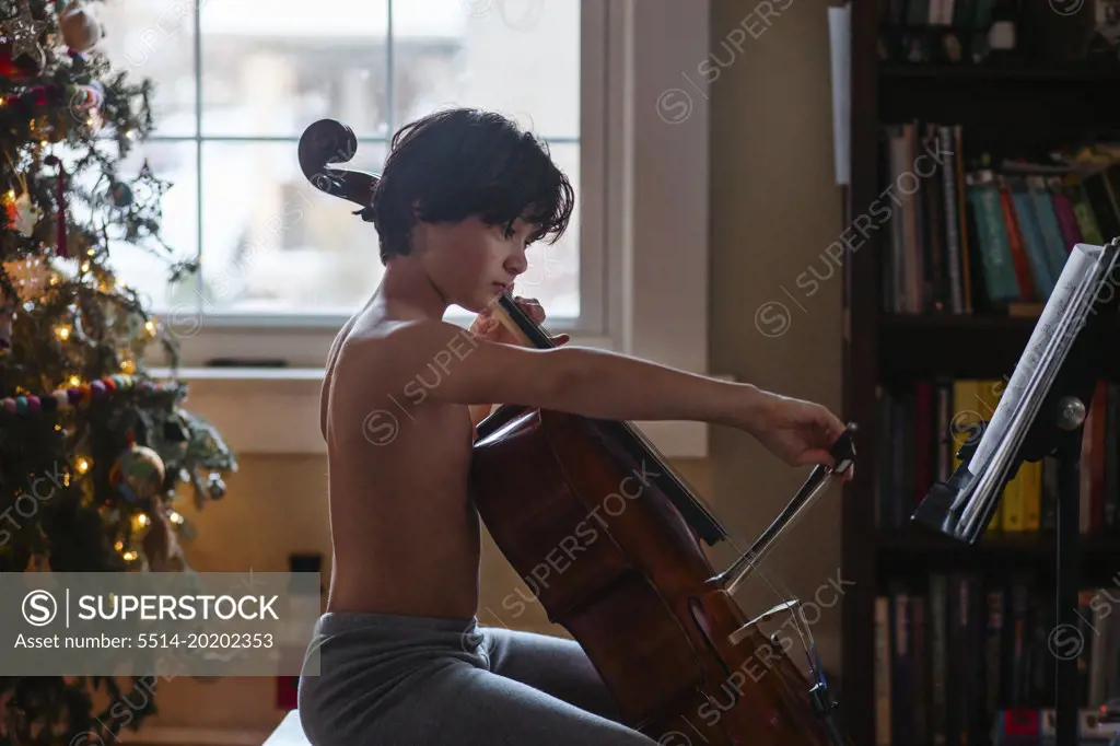 A boy sits by Christmas tree practicing cello by window light