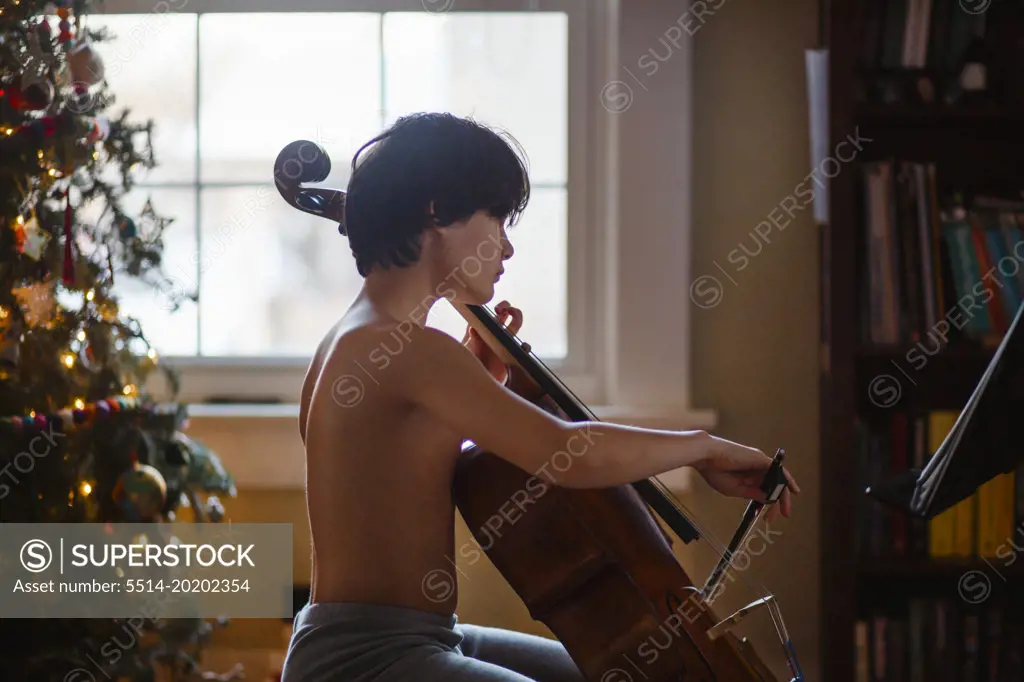 Beautiful boy practices cello by Christmas tree