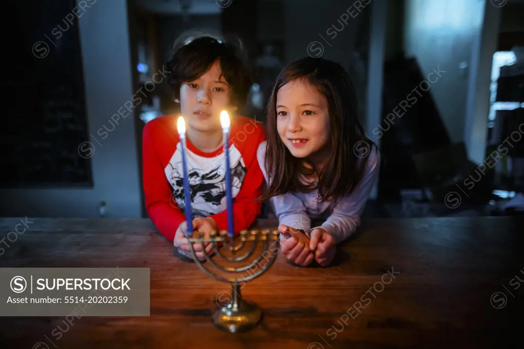 Two children sit at table together looking at lit menorah at Hannukah