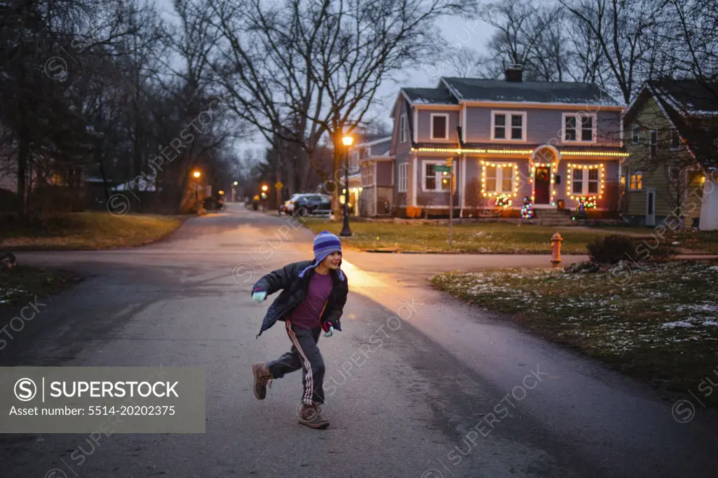 A boy in winter clothes plays on lamplit street at dusk