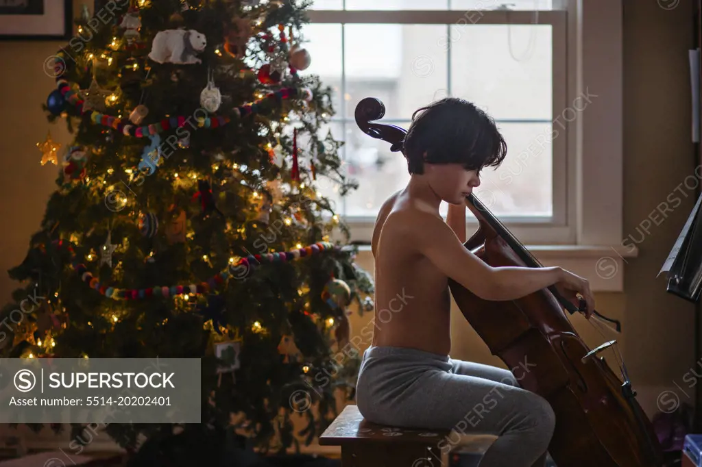 A shirtless boy plays cello by a lit Christmas tree at home