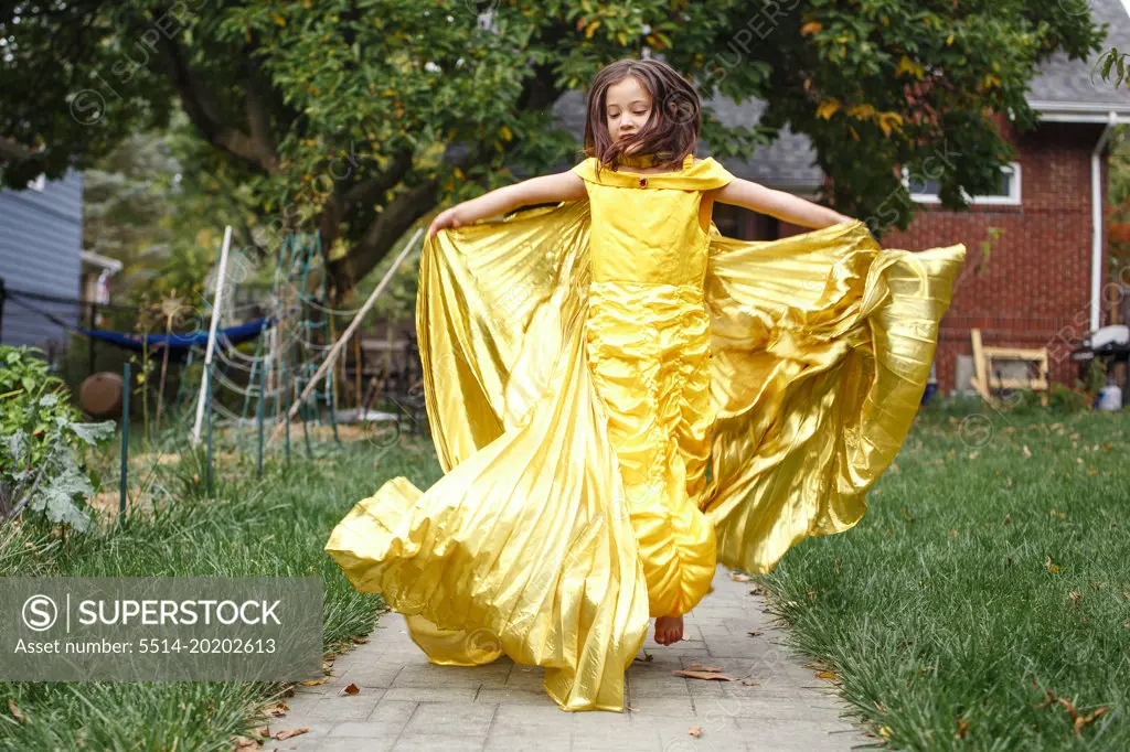 A girl in gold dress and wings leaps barefoot into air in backyard