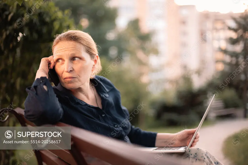 Mature business woman holding laptop talking on mobile phone in park