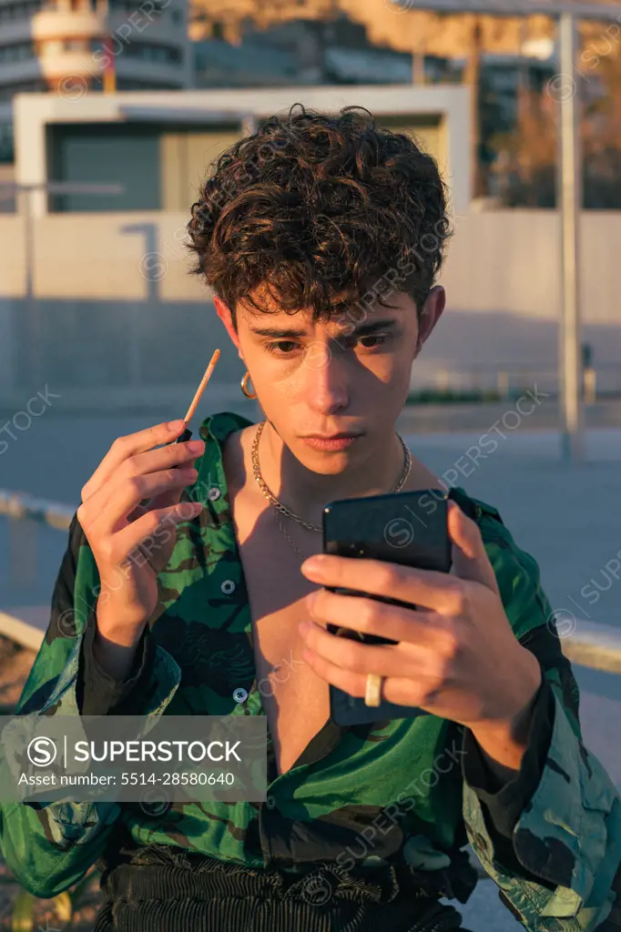Young man wears makeup while watching himself on a phone