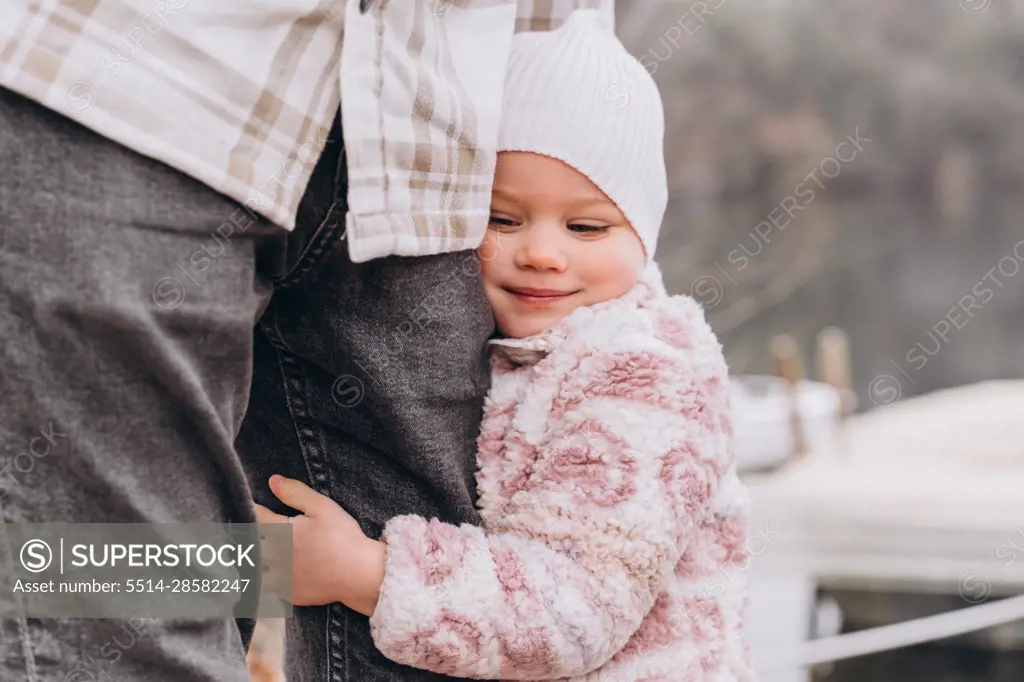 Baby girl hugs dad by the leg in the spring near the boats