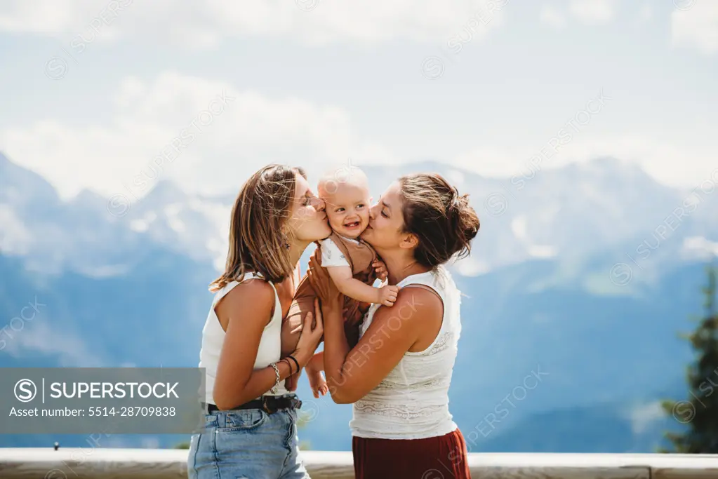 Two moms kissing smiley baby on cheek in summer in mountains