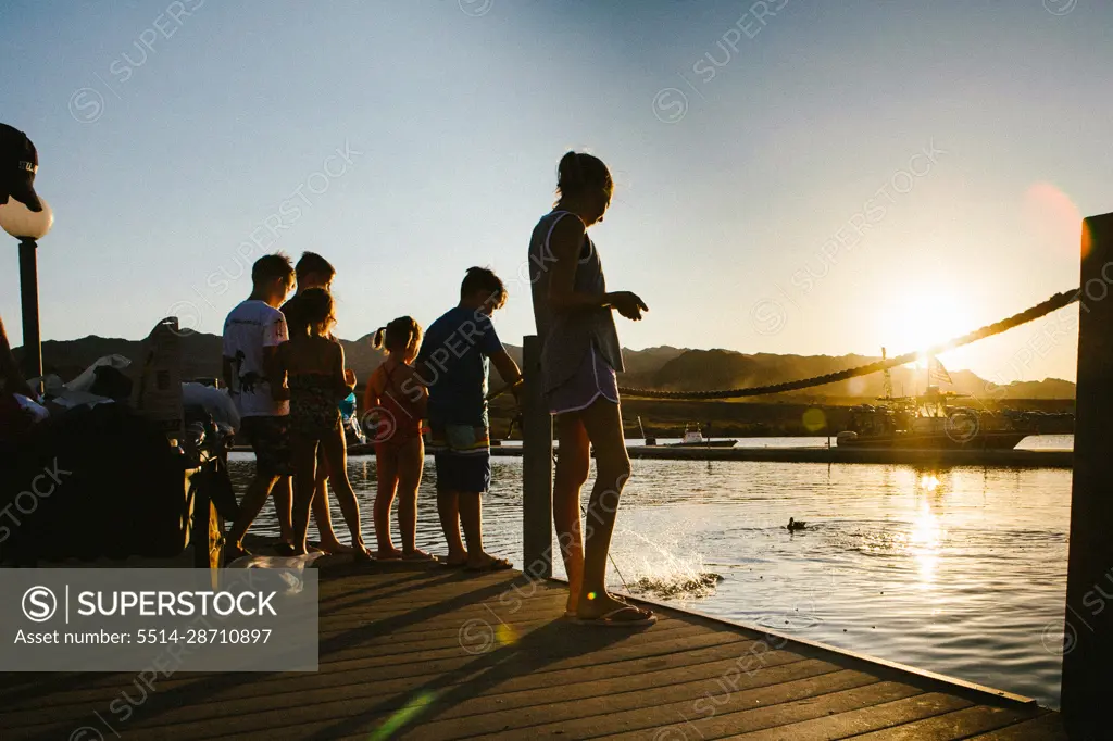 Group of kids stand on dock by lake in summer sunshine and feed fish