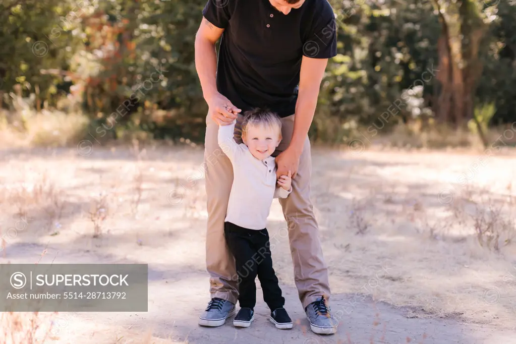 Young dad standing, toddler between legs, holding hands