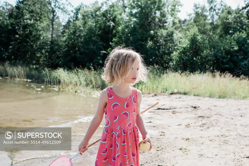 young blonde girl holding a fishing net & apple at the beach in summer -  SuperStock