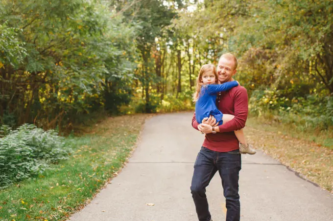 A smiling father holds small daughter in his arms on a tree-lined path