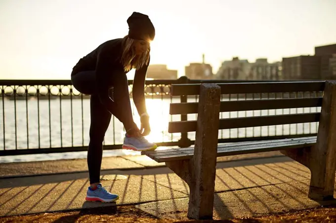 A backlit athletic woman ties her sneaker on a park bench.