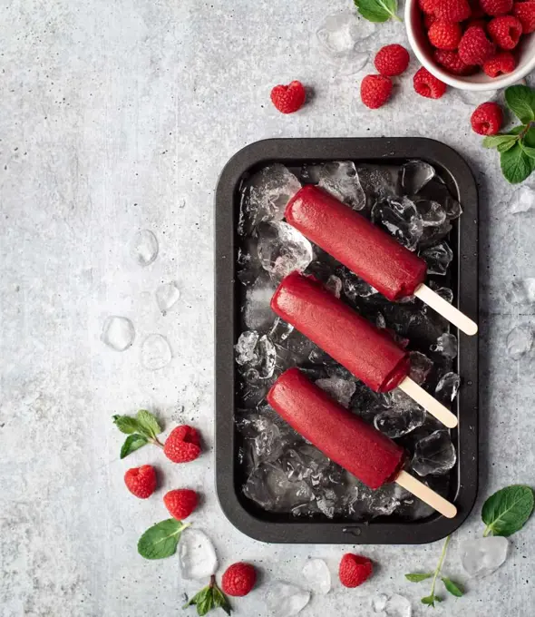Raspberry popsicles on a tray with ice and fruit on grey background.
