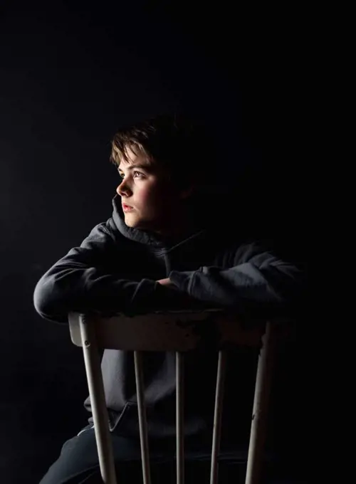 Low key portrait of adolescent boy sitting on a chair in a dark room.
