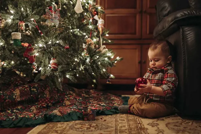 Toddler boy sitting under Christmas tree holding ornament