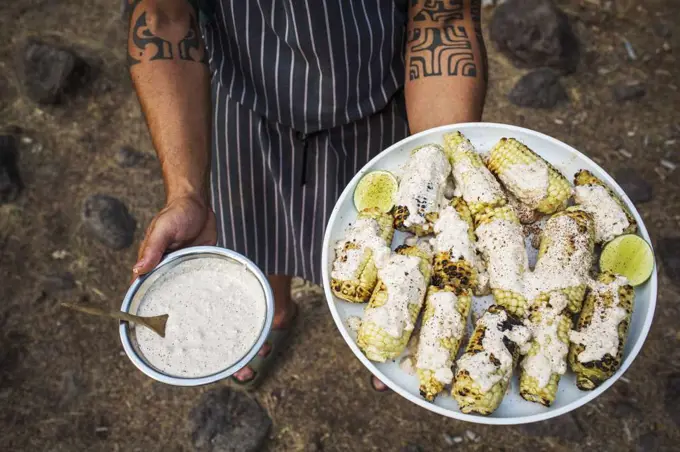 A man with tattooed arms holds a plate of grilled corn and cream