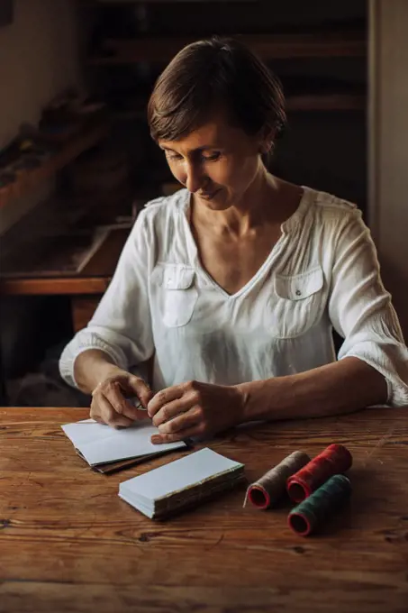 Craftswoman stitching sheets of paper for book, sitting at table