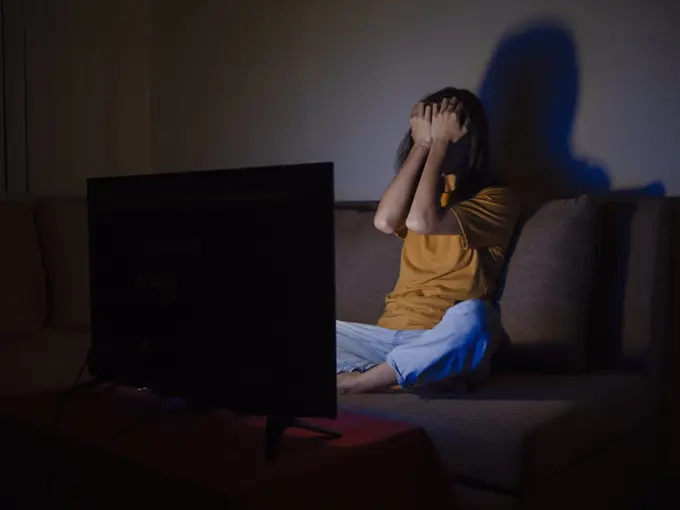 Young woman covering face with palms watching tv at night time