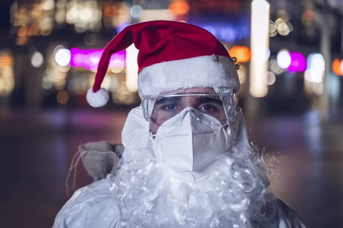 Portrait of a man, with the hat and beard of Santa Claus, but with a face mask, glasses and coverall, to protect himself from Covid 19. In the background the empty city.
