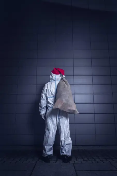 Back View of a volunteer, dressed in the coverall used against coronavirus, the face mask, and the beard and hat of Santa Claus, standing in front of the wall of a hospital, ready to enter to