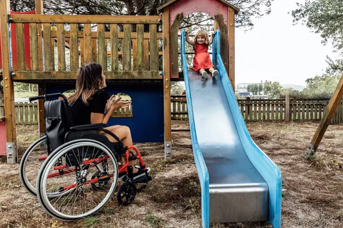 Smiling mother in a wheelchair playing with her little daughter on a s