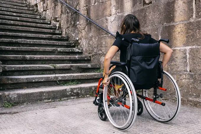 Rear view of a young woman in a wheelchair looking at some stairs. Con