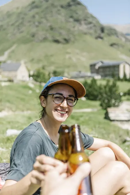 Traveler woman toasting with beer with anonymous man while sitting by the side of a lake in the mountains