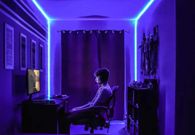 Teenage boy playing video game at desk in room with neon LED lighting.