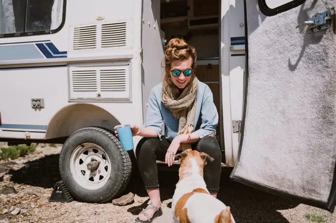 A stylish young woman and her dog sit outside their trailer in the sun