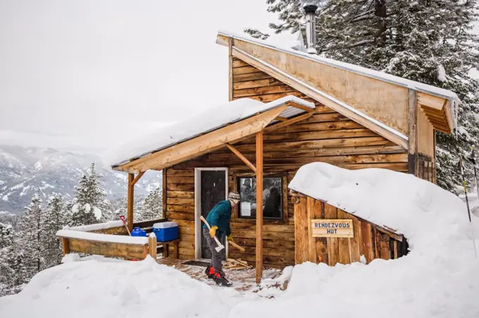 Man chops wood outside of a ski cabin surrounded by deep snow