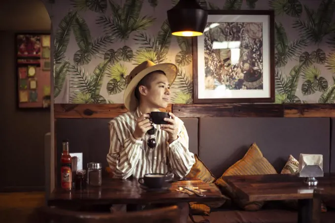 Alternative androgynous young traveller in hat drinks coffee at cafe