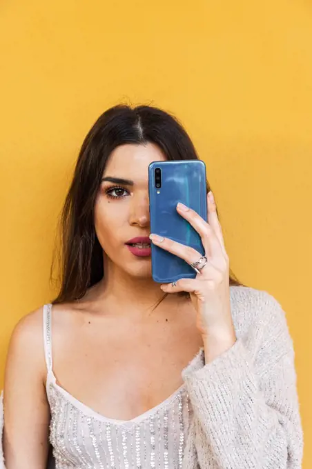 Portrait of Spanish brunette girl covering one eye with her mobile phone on yellow background wall.