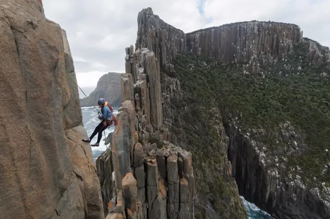 Female rockclimber looks down as she uses a rope to rappel from the edge of a sea cliff to continue exploring the dolerite columns of Cape Raoul, Tasmania, Australia.