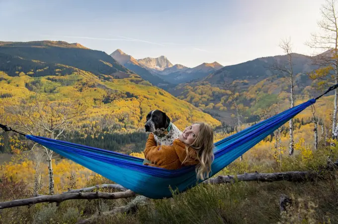 Happy young woman with dog relaxing on hammock in forest during vacation