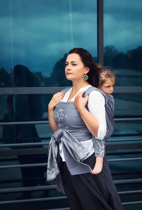 Mother and baby girl sit back in grey wrap sling stay near urban glass