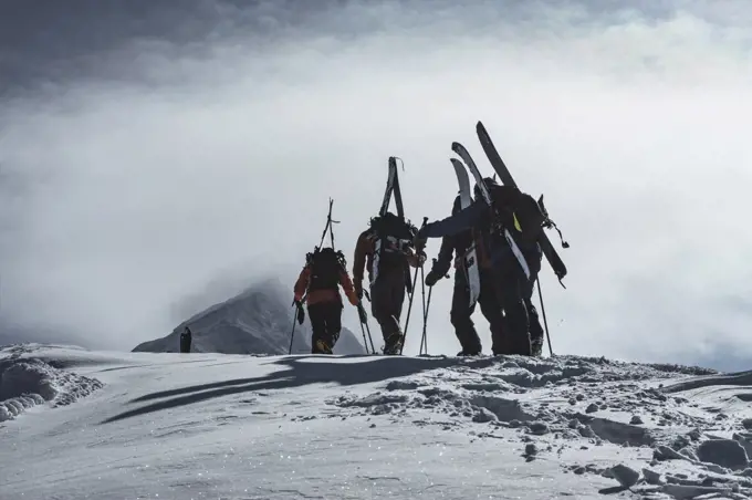 Rear view of people with splitboards and backpacks walking on snow covered mountain peak against cloudy sky