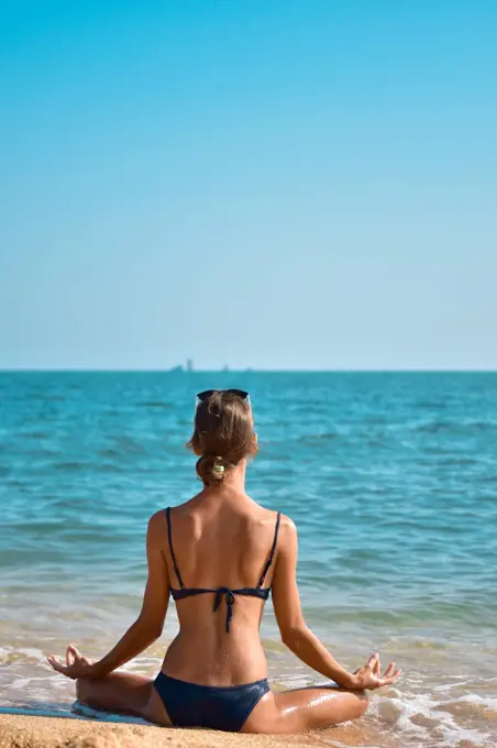 Young woman in a swimsuit sits in a meditative pose on the seashore