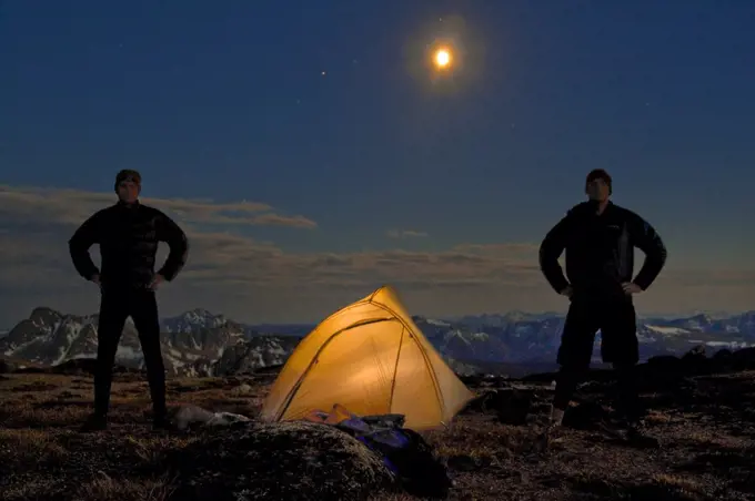 Two men stand next to their lit up tent on a mountain summit.