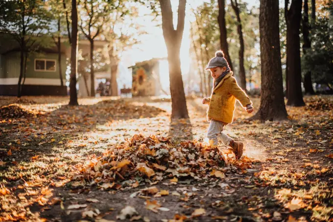 Stylish boy having fun in autumn city park. Happy kid walking among fallen leaves. Kids fashion. Boy wearing trendy yellow coat, cap and scarf. Smiling young boy outdoors. Kid jumping and run