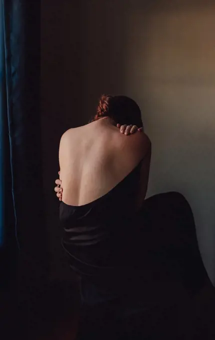Back view of sad woman bent forward with back exposed in a dark room.