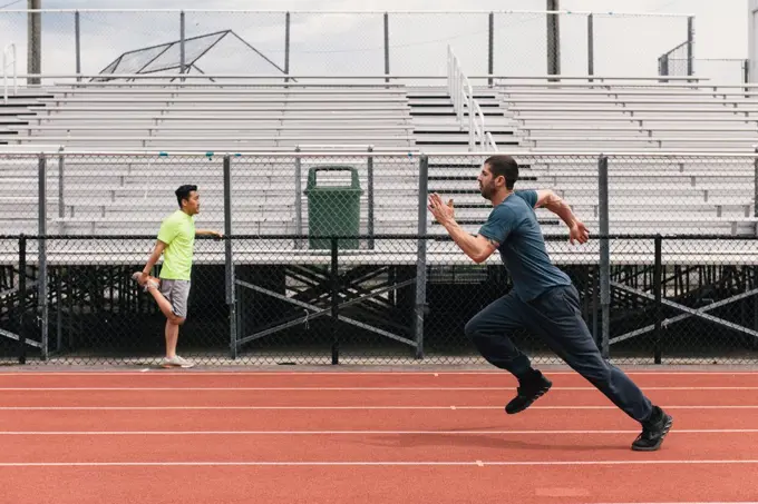 Man running in spring down track while friend stretches by bleachers