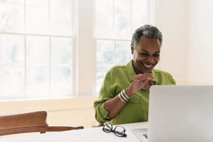 Woman smiling and waving at her laptop while seated at home table