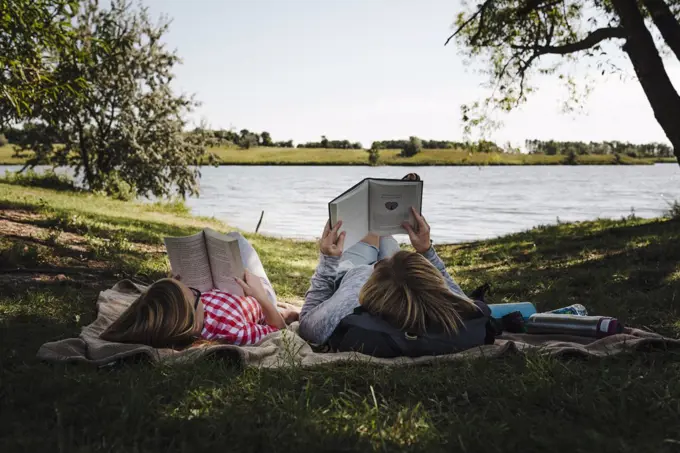 Mom and daughter read on a blanket beside lake on a sunny summer day