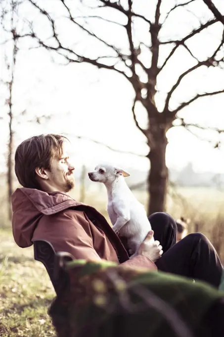 Happy young man with jacket sits outdoors with dog on lap in sunshine