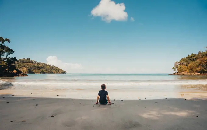 Boy sitting alone on tropical beach looking at the ocean on sunny day.