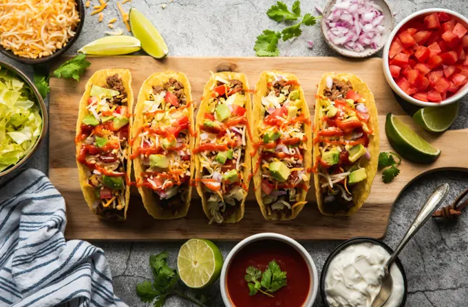 Hard shell tacos on wooden board with beef, lettuce, tomatoes, cheese.